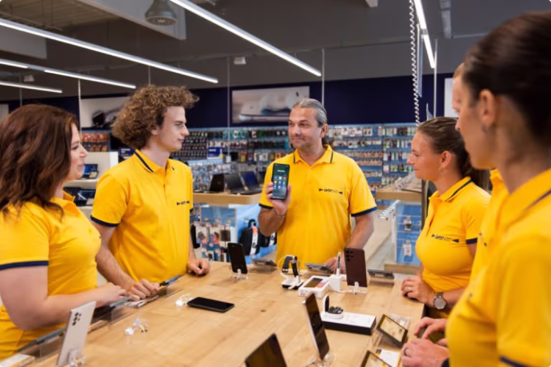 Group of Datart employees around the table looking at another Datart employee who is showing them a new smartphone